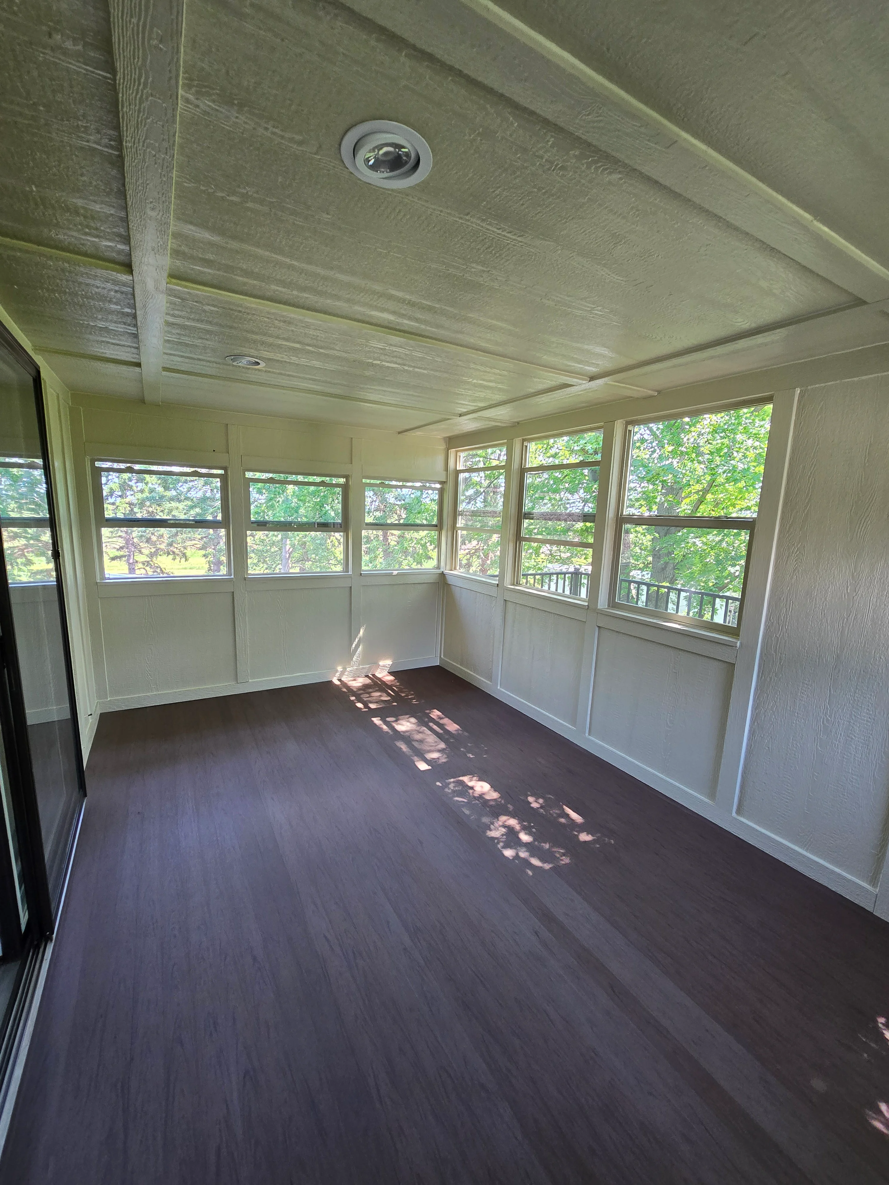 Bright sunroom with dark hardwood floors, white wainscoting, and multi-pane windows overlooking trees